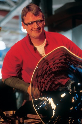 Man with short dark hair wearing safety glasses works a large piece of glass on the end of a punty.