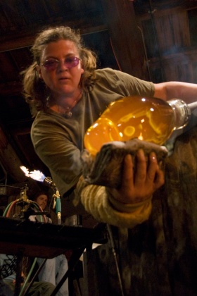 Woman wearing purple glasses with her hair in pigtails shapes a piece of hot glass with newspaper.